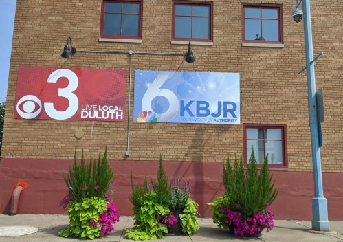 Signage for CBS 3 and KBJR 6 on a brick building, with colorful flower planters in front.
