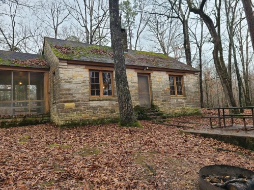 A rustic stone cabin surrounded by trees and fallen leaves, with a small fire pit in the foreground.