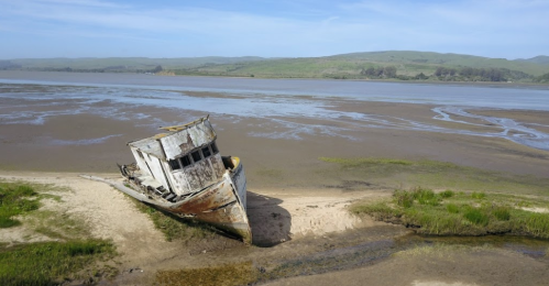 An abandoned, weathered boat rests on a sandy shore, surrounded by calm waters and green hills in the background.