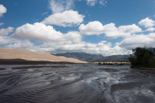 A scenic view of sand dunes under a blue sky with clouds, mountains in the background, and a wet, reflective foreground.