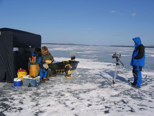 Two people on an icy lake, one sitting by a shelter and the other filming with a camera. Snow and ice cover the ground.