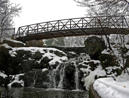 A wooden bridge spans a rocky waterfall, surrounded by snow-covered trees and a serene winter landscape.