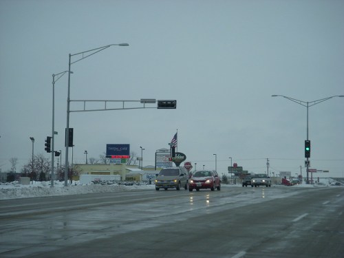 A snowy street scene with traffic lights, cars, and a sign in a commercial area under a gray sky.