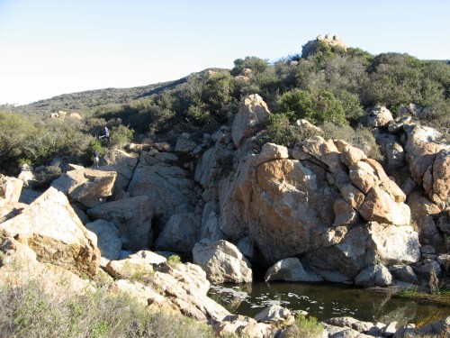Rocky landscape with large boulders and a small pond, surrounded by greenery and hills under a clear blue sky.
