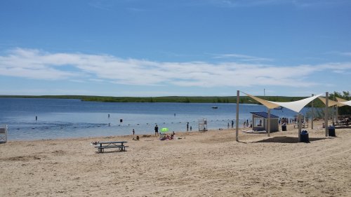 A sunny beach scene with people swimming, a picnic table, and shaded areas near a calm lake and green hills in the background.