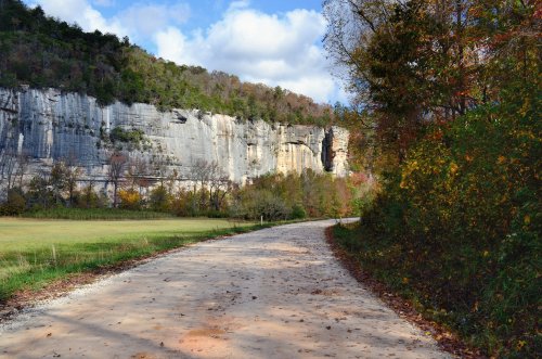 A winding dirt road leads through a scenic landscape with a towering cliff and colorful autumn foliage.