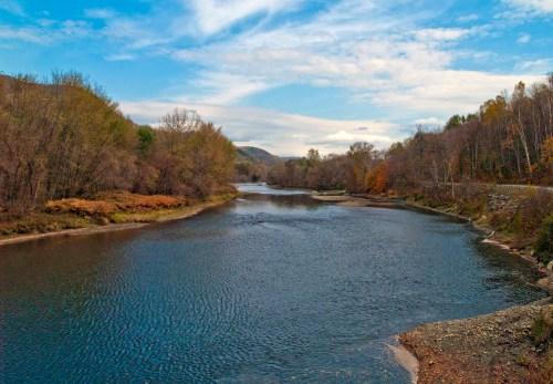 A serene river flows through a landscape of trees with autumn colors under a blue sky with scattered clouds.