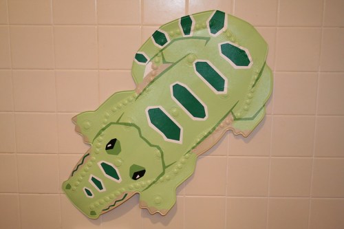 A green crocodile-shaped bath mat with black and white accents, placed on a tiled bathroom wall.