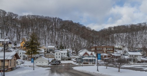 Snow-covered houses nestled at the base of a wooded hill, with a cloudy sky overhead.