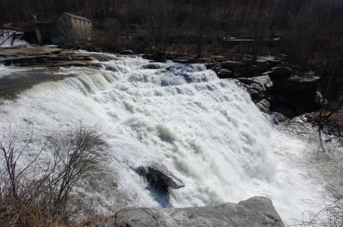 A powerful waterfall cascades over rocky ledges, surrounded by bare trees and a distant building.