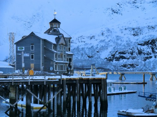 A snowy harbor scene featuring a wooden building on a pier, surrounded by mountains and calm water.