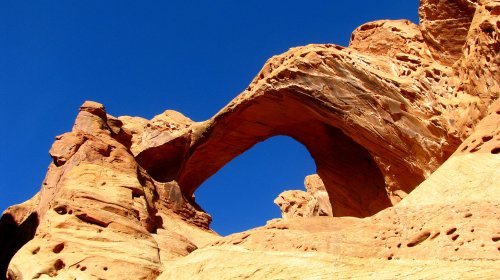 A natural rock arch stands against a clear blue sky, surrounded by textured red sandstone formations.