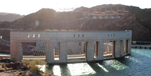 A large concrete dam structure with multiple openings, surrounded by rocky terrain and water reflecting sunlight.