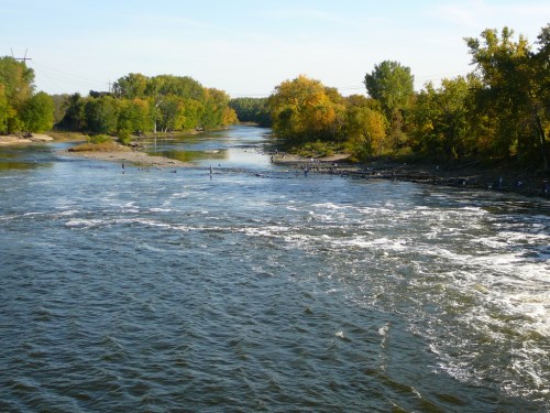 A serene river flows through a landscape of trees with autumn foliage under a clear blue sky.