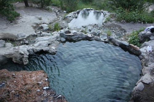 A natural hot spring surrounded by rocks and greenery, with clear water reflecting the surroundings.