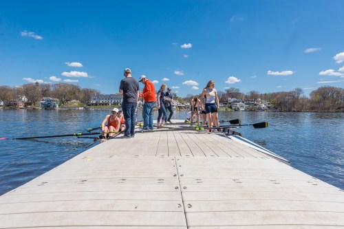 A group of people on a dock by a river, preparing for rowing activities on a sunny day with blue skies.