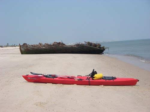 A red kayak rests on a sandy beach near a weathered shipwreck in calm waters.