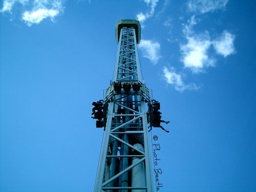 A tall amusement park drop tower against a blue sky, with riders suspended near the top.