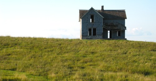 An abandoned, weathered house sits on a grassy hill under a clear blue sky.