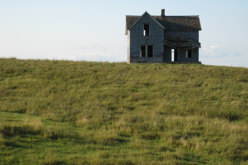 Abandoned blue house on a grassy hill under a clear sky, with broken windows and a weathered exterior.