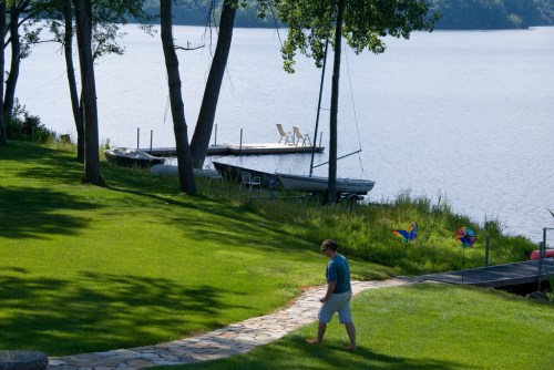 A person walks along a stone path by a lake, with boats and chairs visible on the shore. Lush green grass surrounds the area.