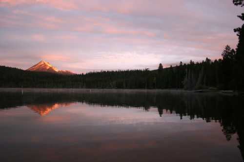 A serene lake reflects a mountain at sunrise, surrounded by trees and a colorful sky.