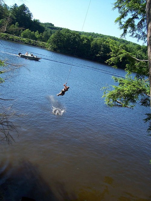 A person swings from a rope into a river on a sunny day, with a boat in the background.