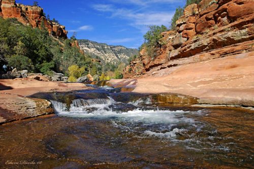 A serene river flows over smooth rocks, surrounded by red cliffs and lush greenery under a clear blue sky.