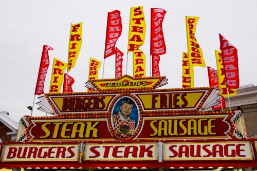 Colorful food stand sign featuring burgers, fries, steak, and sausage with vibrant flags above.