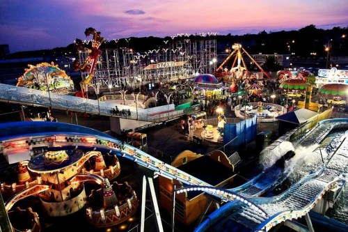 A vibrant amusement park at dusk, featuring rides, games, and colorful lights against a sunset sky.