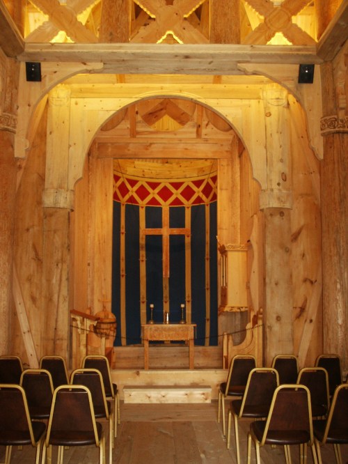 Interior of a wooden structure featuring a simple altar, decorative archways, and rows of empty chairs.