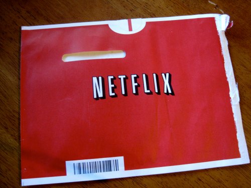 A red Netflix envelope with a cut-out handle and a barcode at the bottom, resting on a wooden surface.