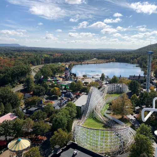 Aerial view of a theme park with a roller coaster, lake, and colorful attractions surrounded by trees and hills.