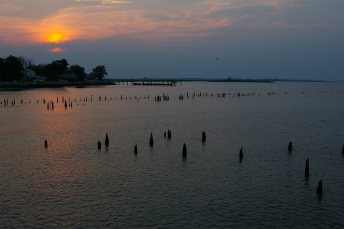 Sunset over a calm water body, with wooden pilings emerging from the water and a distant pier along the shore.