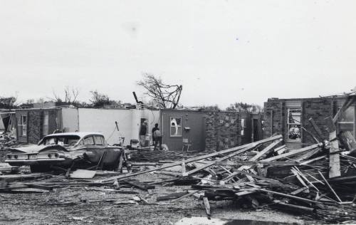 A black-and-white photo of a tornado-damaged neighborhood with debris, a wrecked car, and a person inspecting the ruins.