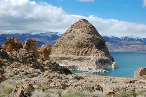 A rocky formation rises from a turquoise lake, surrounded by mountains and a partly cloudy sky.