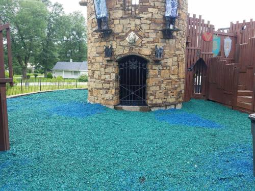 A stone castle play structure with a gated entrance, surrounded by blue and green rubberized ground cover.