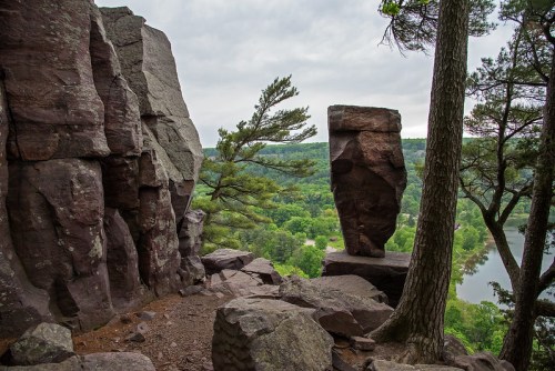 A rocky landscape featuring a tall, balanced rock formation surrounded by trees and a view of a river below.