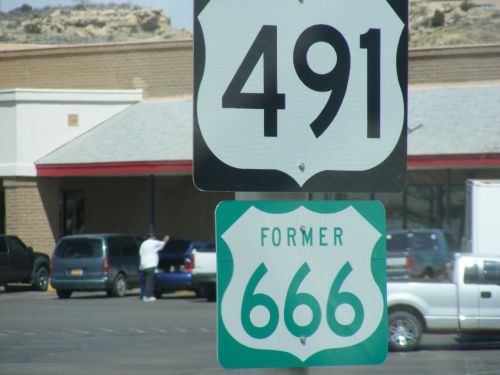A highway sign displaying Route 491 above a sign for the former Route 666, set in a parking lot.