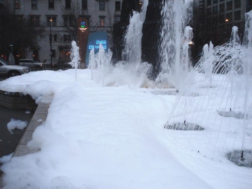 A fountain surrounded by foam, with a digital clock displaying the time in a city setting.