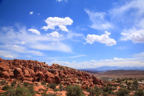 A scenic view of red rock formations under a blue sky with fluffy clouds and distant mountains.