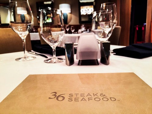Table setting at a restaurant featuring a menu for "36 Steak & Seafood," with glasses and elegant decor.