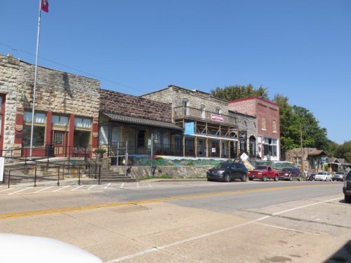 Historic stone buildings line a street, featuring shops and a clear blue sky above. Cars are parked along the road.