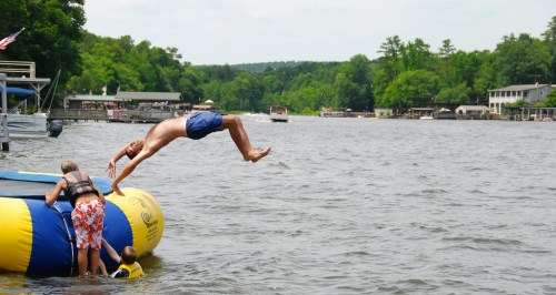A person jumps off a trampoline into a lake while others play nearby, surrounded by trees and boats.