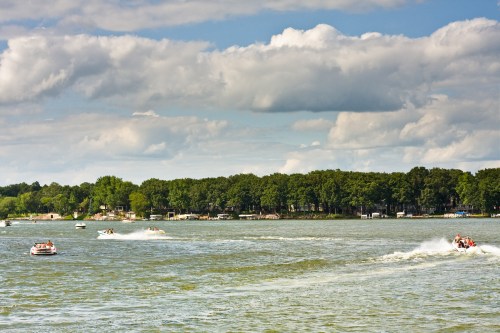 Boats speeding across a lake with a tree-lined shore under a partly cloudy sky.
