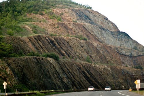 A rocky hillside with layered geological formations alongside a highway, with cars driving on the road below.