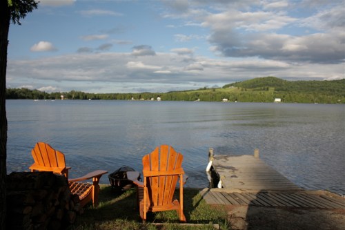 Two orange Adirondack chairs by a calm lake, with a wooden dock extending into the water and green hills in the background.
