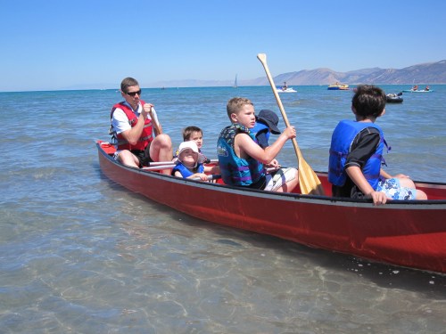 A group of children and an adult paddling a canoe on a sunny day in shallow water.