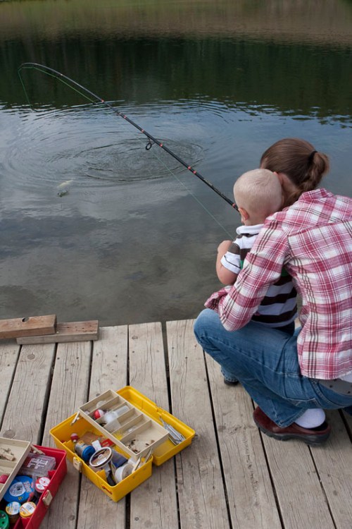 A person and a child fish from a wooden dock, with a tackle box nearby and ripples in the water from a caught fish.