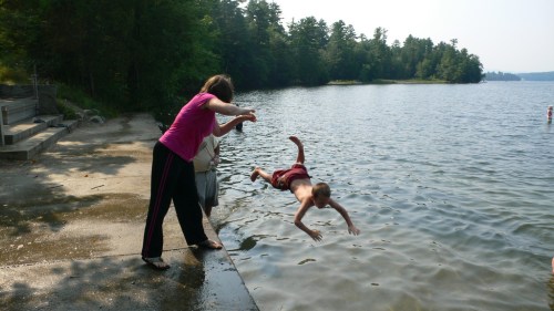 A child jumps into a lake while an adult encourages them from the shore on a sunny day. Trees line the background.